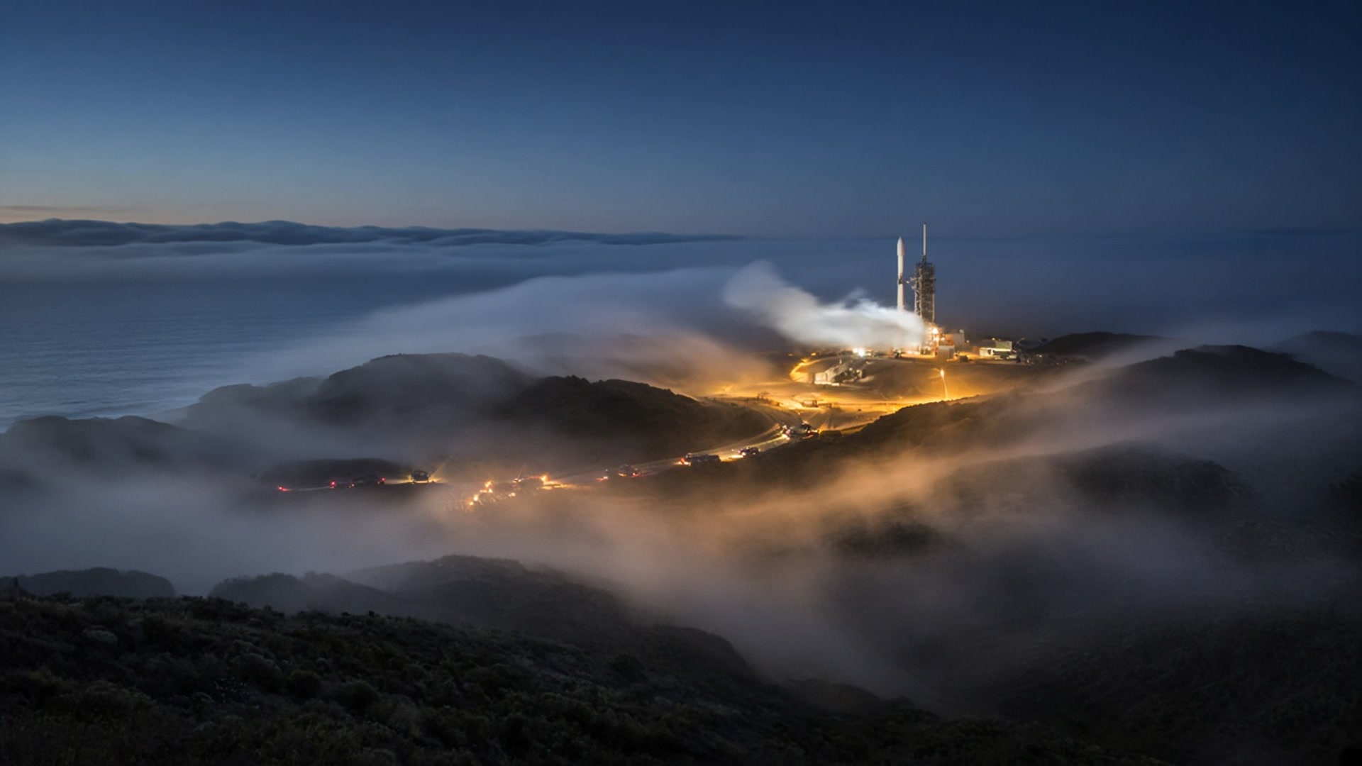 Before dawn on the California coast, fog rolls past a floodlit launch pad where a rocket stands venting.