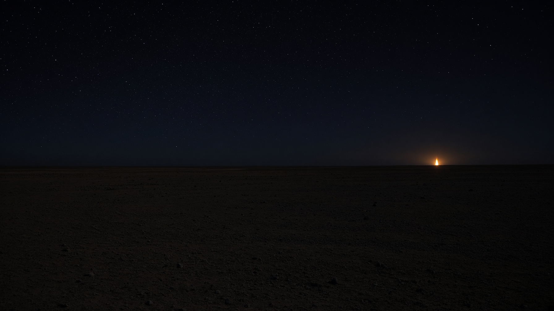 A distant gas flare on a dark industrial horizon, black sky, empty ground.